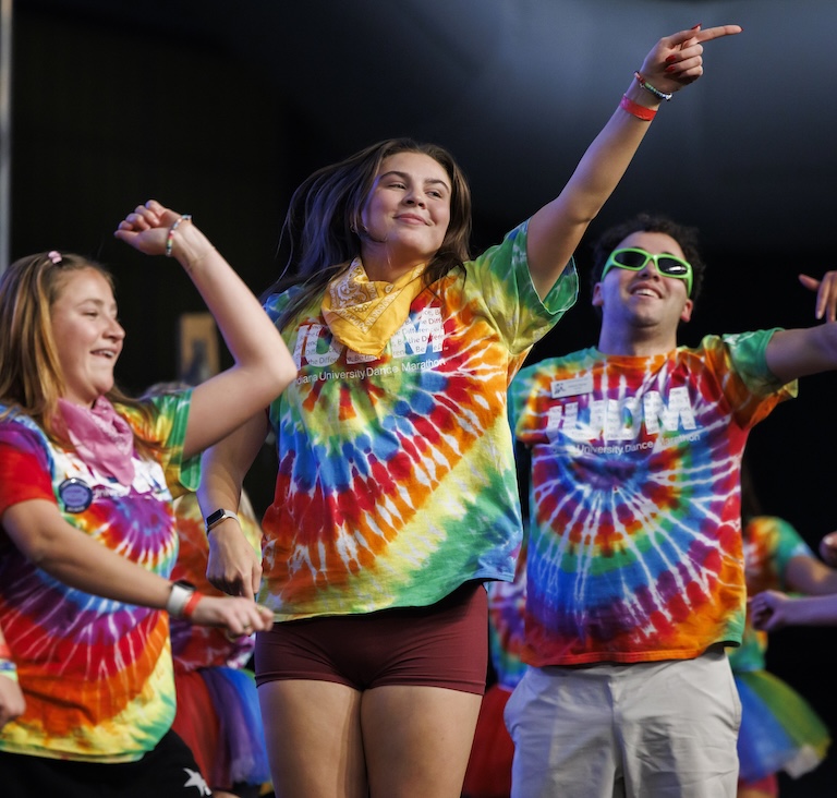 three students on a stage wearing tie-dye shirts with the IU Dance Marathon logo on them